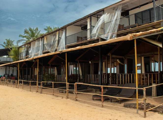 A beach house featuring a balcony and a wooden fence, surrounded by sandy shores and a clear blue sky.