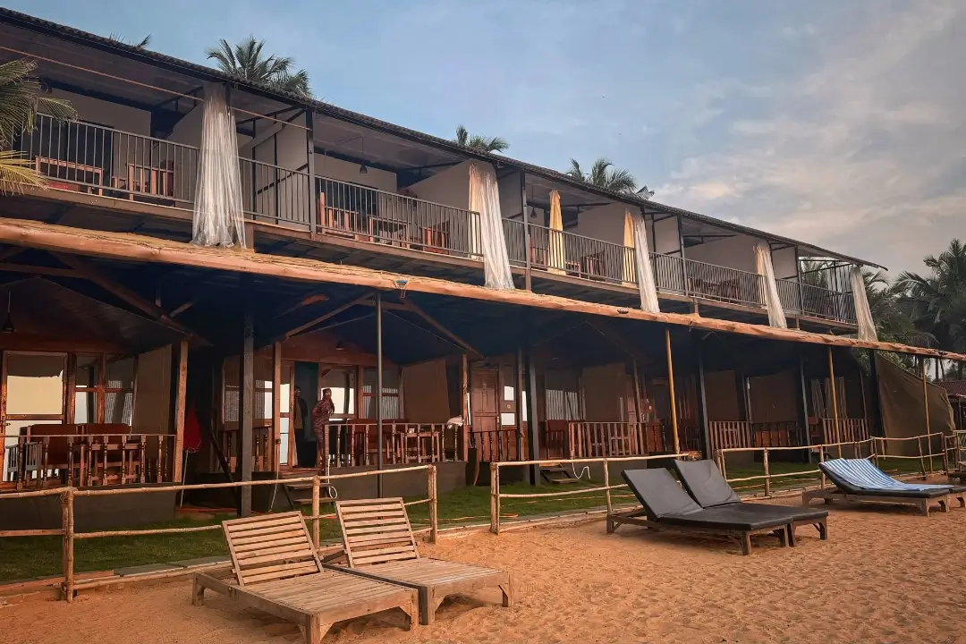 A beach house featuring a balcony and a wooden fence, surrounded by sandy shores and a clear blue sky.