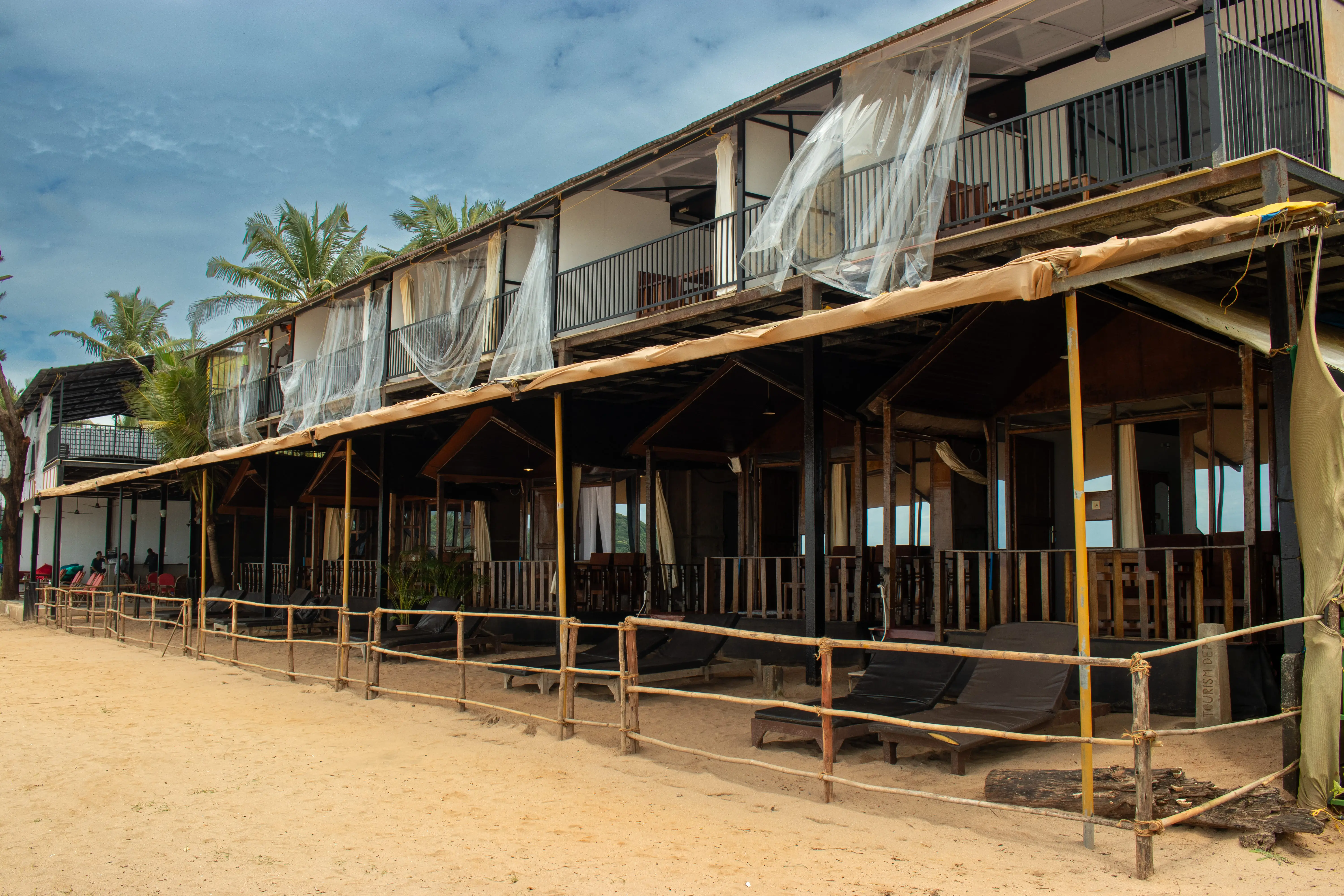 A beach house featuring a balcony and a wooden fence, surrounded by sandy shores and a clear blue sky.